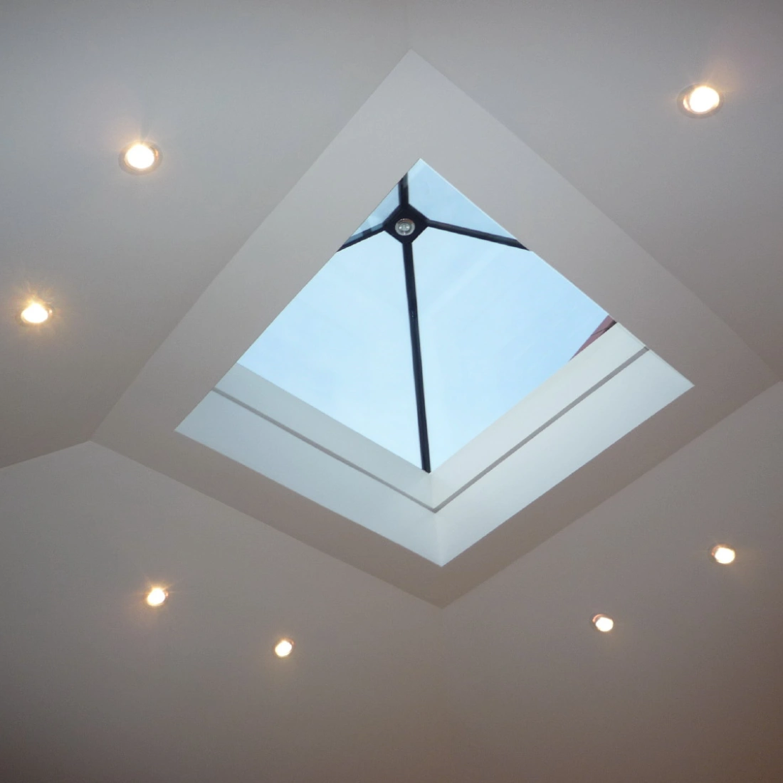 Skylight in a ceiling, surrounded by recessed lights, with a clear blue sky visible through the glass.