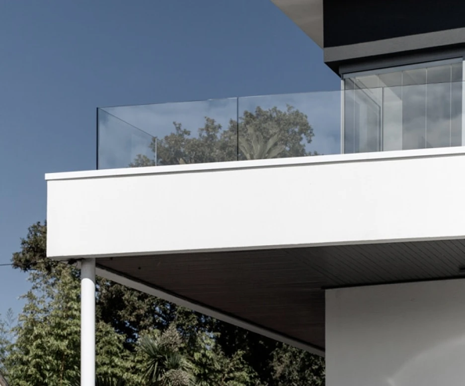 Fachada moderna de una casa con un balcón de vidrio, rodeada de vegetación y un cielo azul.