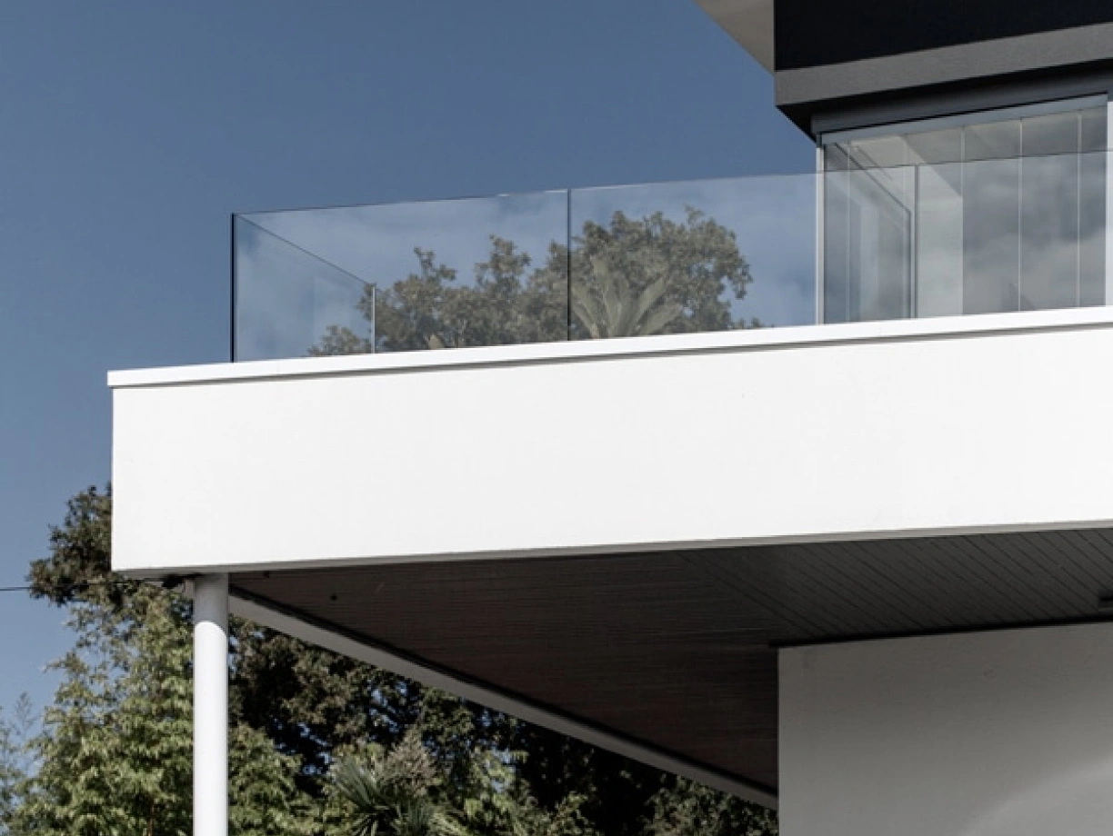 Fachada moderna de una casa con un balcón de vidrio, rodeada de vegetación y un cielo azul.