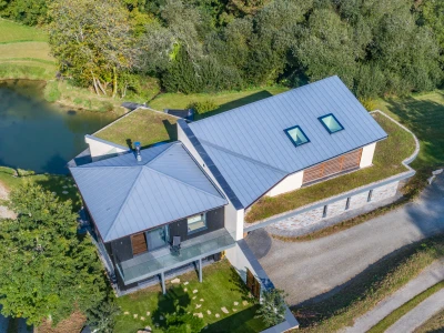 Moderne Architektur eines Hauses mit einem schrägen Dach, umgeben von Bäumen und einem kleinen Teich. Eine geschwungene Auffahrt führt zum Eingang des Hauses.