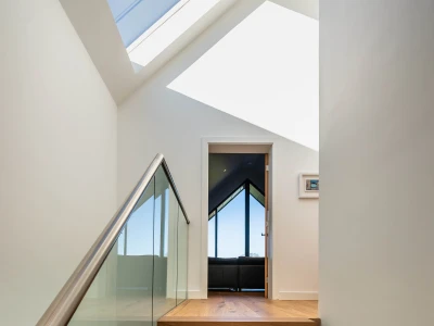 Escalera que lleva a un pasillo iluminado por un tragaluz. Al fondo, una puerta abierta revela una sala con ventanas grandes que ofrecen vistas al exterior.