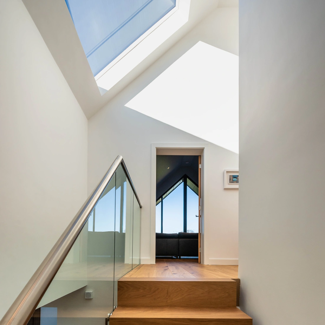 Escalera que lleva a un pasillo iluminado por un tragaluz. Al fondo, una puerta abierta revela una sala con ventanas grandes que ofrecen vistas al exterior.