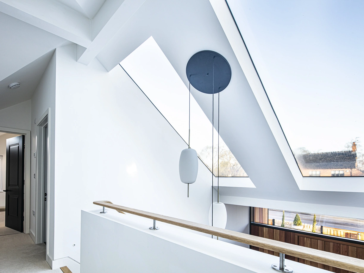 Bright interior with white walls and a high ceiling featuring large skylights. A wooden railing overlooks a lower level, with modern light fixtures hanging from the ceiling. A doorway leads to another room, and views of an outdoor area are visible through the windows.