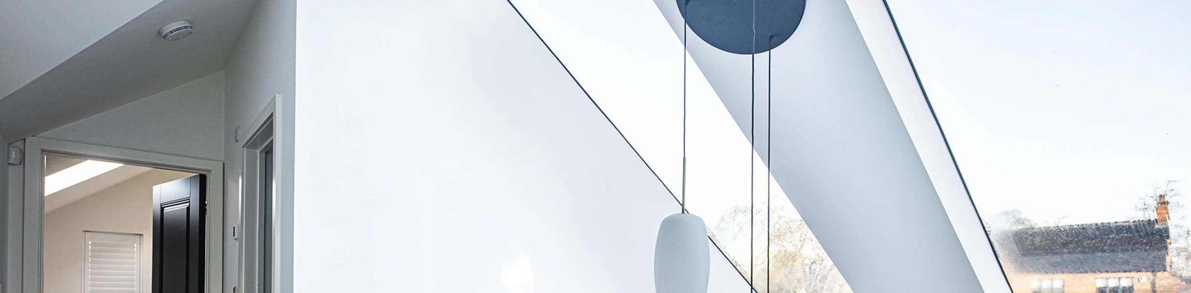 Vue d’un espace intérieur lumineux avec un grand plafond incliné. Des fenêtres situées dans le plafond laissent entrer la lumière naturelle. Un garde-corps en bois sépare l’escalier, et un luminaire suspendu apporte une touche moderne.