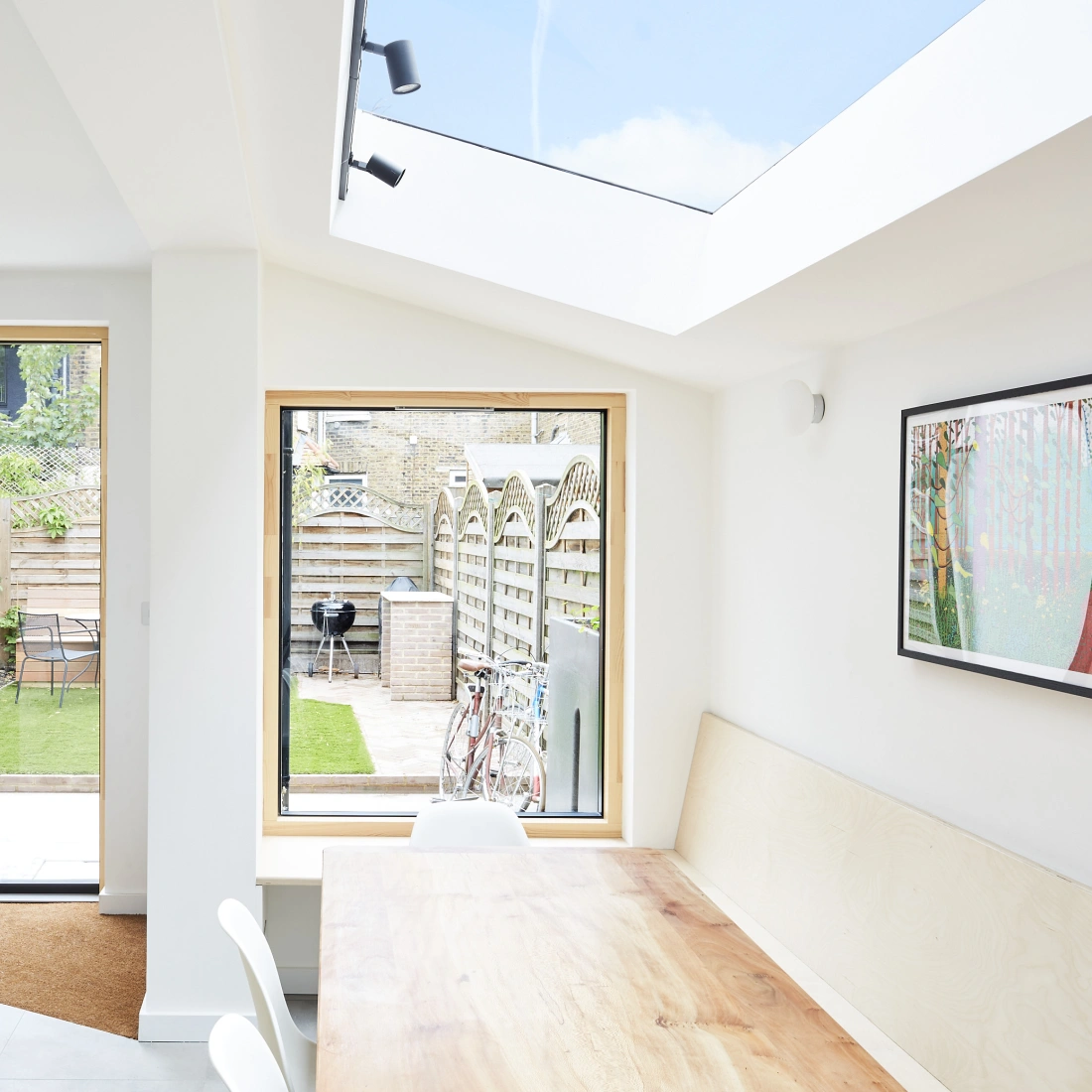 Espacio interior luminoso con una mesa de madera y sillas blancas. Ventanas grandes ofrecen vistas a un jardín con cercas de madera y un mobiliario exterior. Un techo acristalado permite la entrada de luz natural.