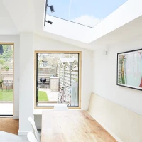 A bright and modern kitchen with a large wooden dining table, featuring a vase of flowers. The room has multiple large windows showcasing a well-maintained garden with a patio and bicycle. A skylight illuminates the space, enhancing the airy atmosphere.