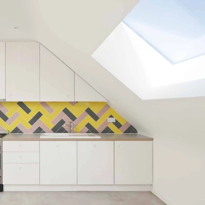 Modern kitchen featuring a sloped ceiling, white cabinetry, and a vibrant yellow, pink, and black herringbone tile backsplash. A stainless steel stove is positioned against the wall, with a skylight above allowing natural light to fill the space.