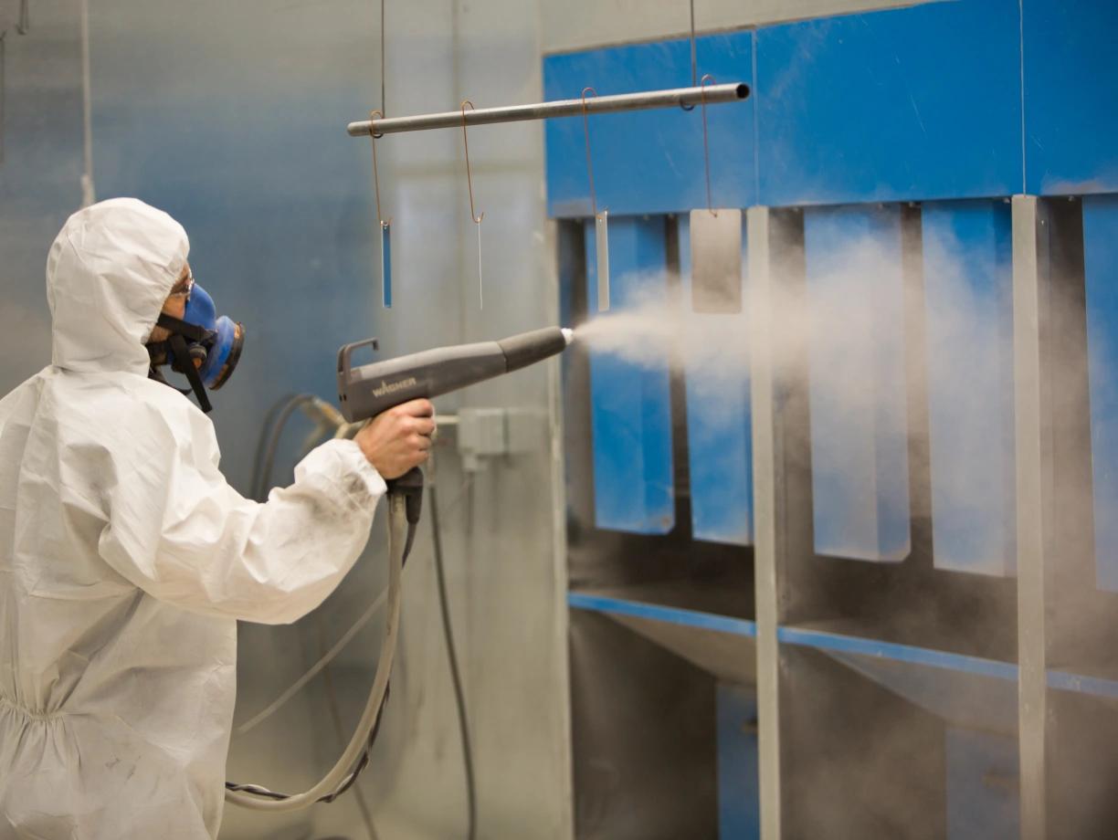 A person in a white protective suit and gas mask is using a spray gun to apply a coating inside a industrial spray booth. Soft clouds of mist are visible around the spray area, and the background features blue panels.