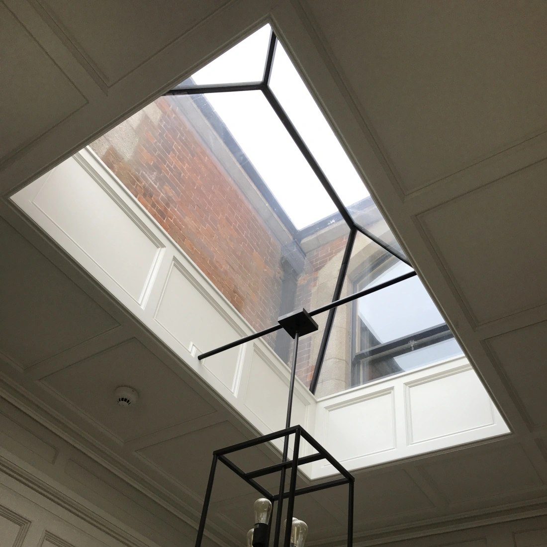 Ventana de tejado rectangular con marco negro, iluminando una habitación con un elegante candelabro de diseño minimalista.