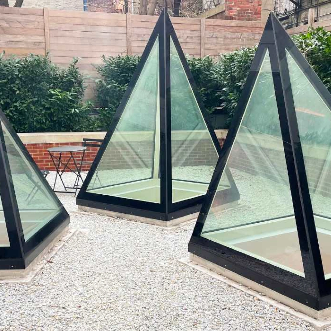 Three glass and black-framed pyramids set in a gravel area surrounded by greenery. The pyramids are arranged at different angles, creating a modern architectural design.
