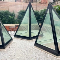 Three glass pyramids with black frames are situated in a gravel area surrounded by lush greenery and a wooden fence. A small black table and chairs are visible nearby.