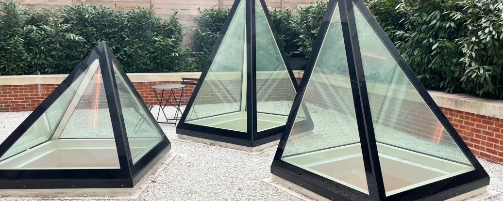 Three pyramid-shaped glass structures sit on a gravel surface, surrounded by low greenery and a brick wall. A small black table is visible nearby.