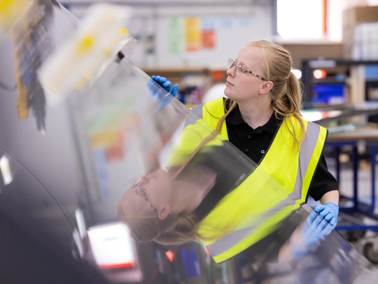 A person wearing a yellow safety vest and blue gloves inspects a shiny, reflective surface in a workshop. The individual has long blonde hair tied back and is focused on their task.