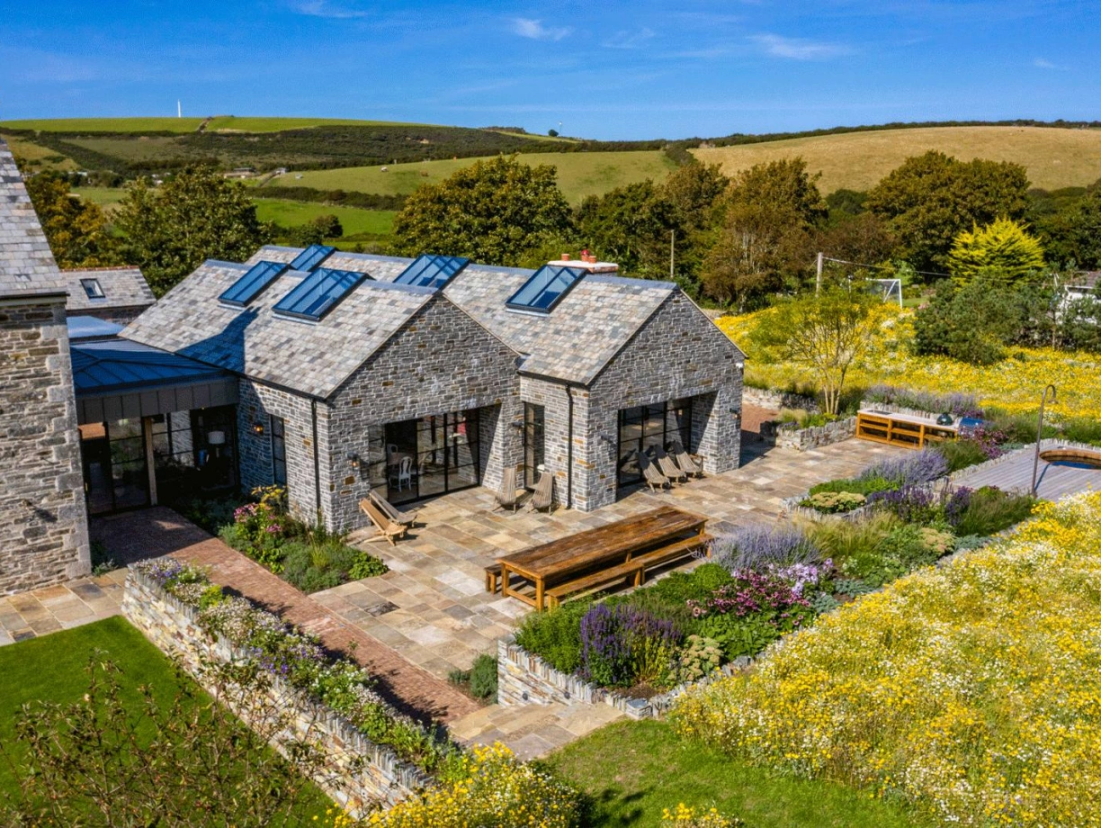 A contemporary stone house with a slate roof sits amidst a vibrant garden filled with yellow flowers. The house features large windows and skylights, while a spacious patio includes a long wooden dining table and benches surrounded by lush greenery. Rolling hills can be seen in the background under a clear blue sky.