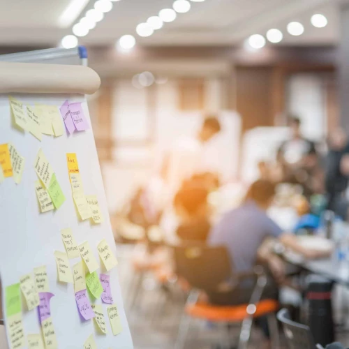 A whiteboard with colorful sticky notes in the foreground, with a blurred group of people engaged in discussion around tables in a well-lit room.