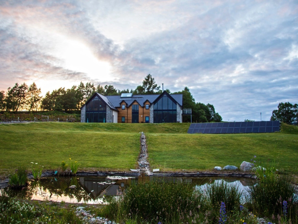 Casa moderna de dos pisos con paneles solares, rodeada de un paisaje verde y un estanque. El cielo está nublado con tonalidades anaranjadas y azules.