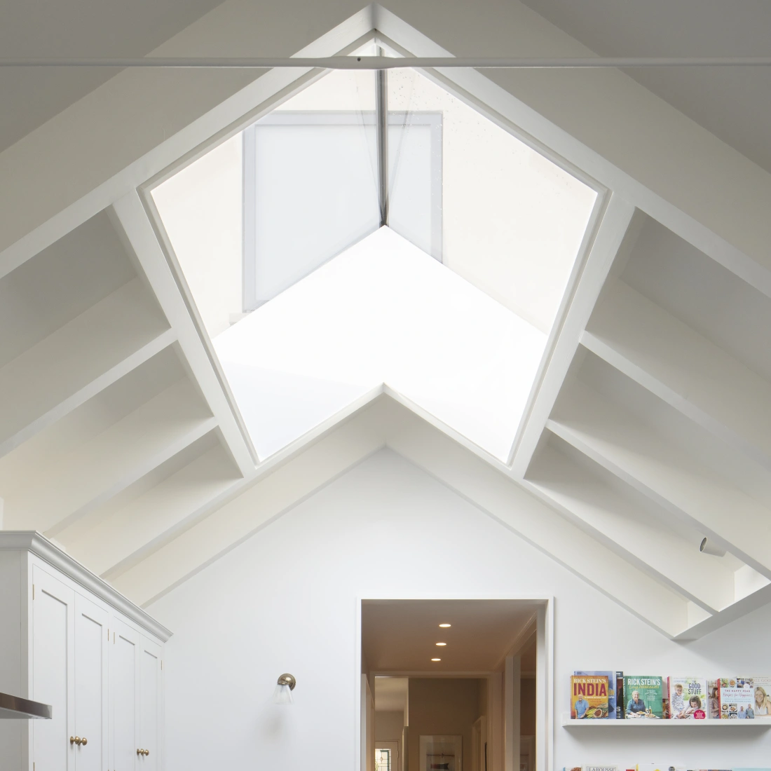 Eine moderne Küche mit einer großen, mehrseitigen Oberlichter und sichtbaren Holzstuhlträgern. Auf der linken Seite befinden sich weiße Küchenschränke mit goldenen Armaturen. Rechts steht ein Regal mit verschiedenen Kochbüchern. Ein heller Flur führt in den Hintergrund.