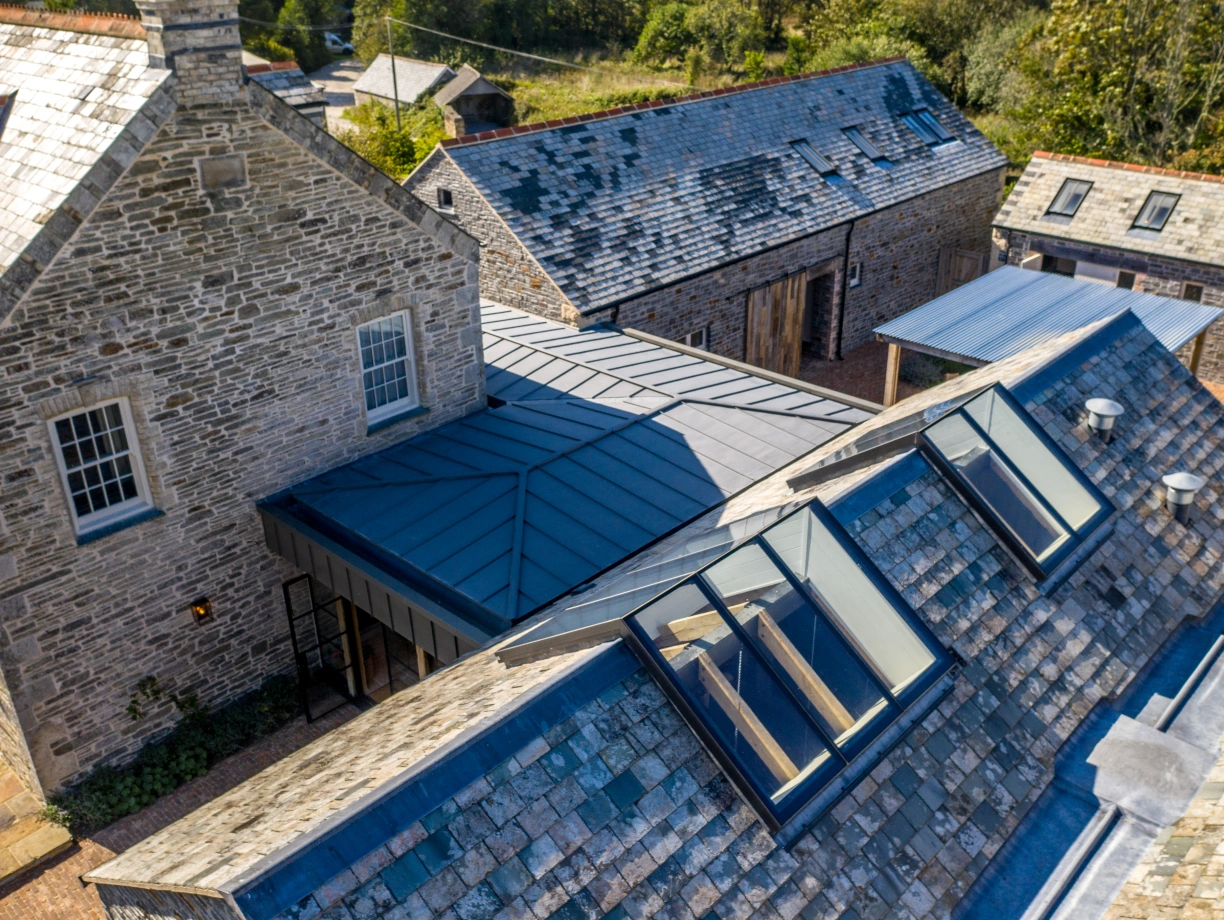 Aerial view of a rustic stone house and several nearby buildings, featuring a slate roof and modern skylights. Surrounding landscape includes trees and open space.