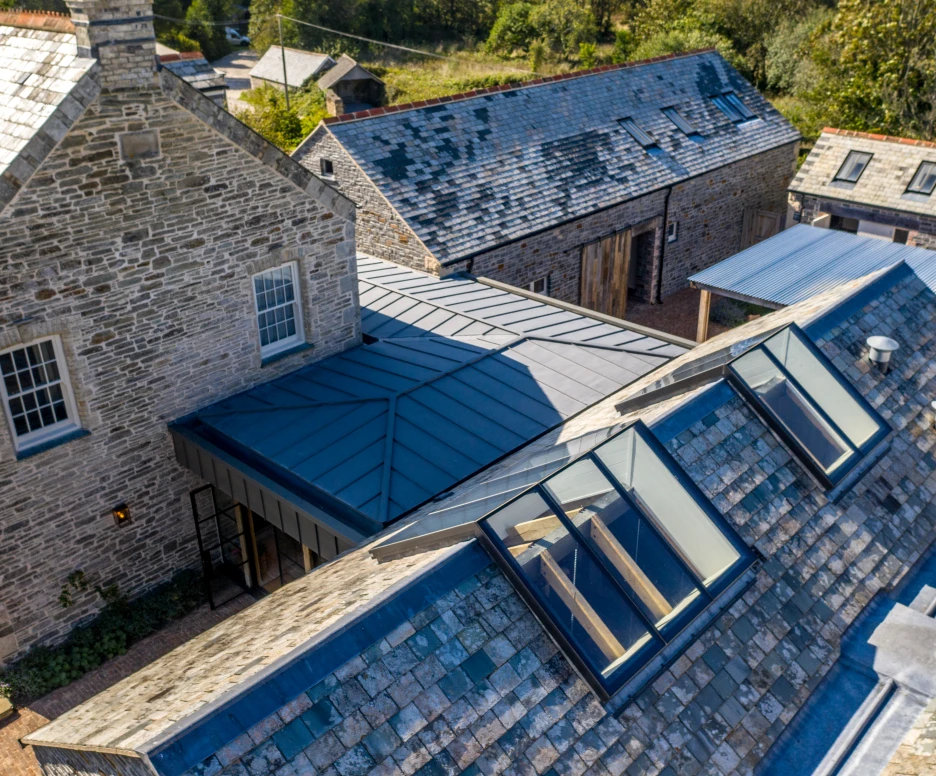 Vue aérienne d'un complexe architectural avec plusieurs bâtiments en pierre et un toit en ardoise. L'un des toits présente des lucarnes modernes, tandis que des entrepôts en bois se trouvent en arrière-plan. La verdure environnante ajoute une touche naturelle.