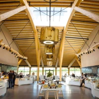 A modern restaurant interior featuring high wooden beams, large skylights, and multiple display counters filled with food. Diners interact in the spacious area, which has a clean, contemporary design.