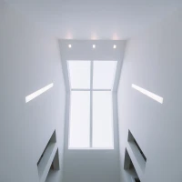 Blick nach oben in einen modern gestalteten Raum mit weißen Wänden und dreieckigen Fenstern, die Tageslicht hereinlassen. An der Decke befinden sich mehrere gleichmäßig verteilte Lichtquellen.