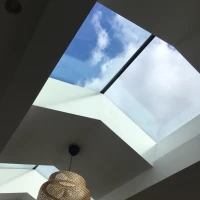 A modern ceiling featuring geometric skylights allowing natural light to flow in. A woven pendant light hangs from the ceiling, contrasting with the white walls and blue sky above.