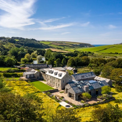 Aerial view of a large stone farmhouse surrounded by lush fields and trees, with rolling hills and the sea visible in the background under a clear blue sky.