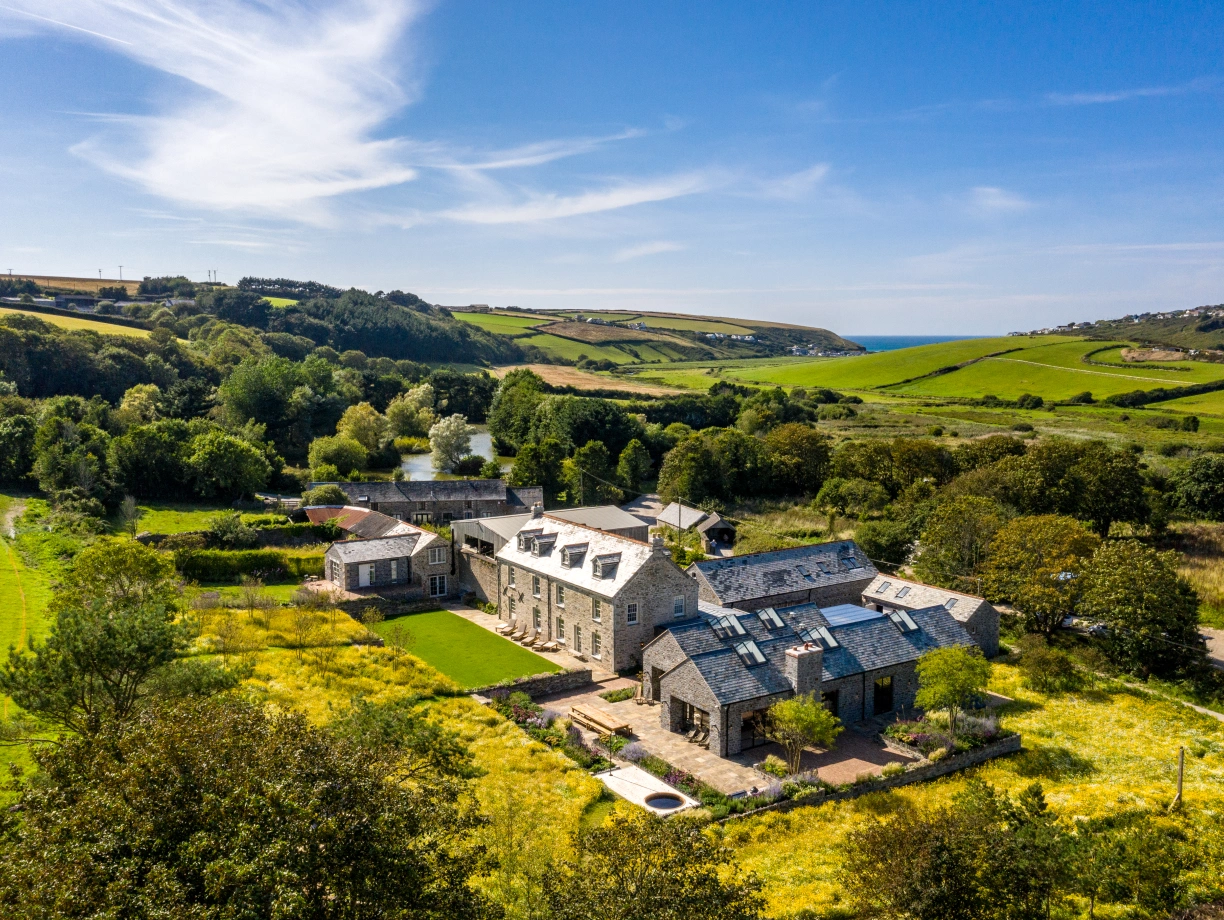 A picturesque countryside scene featuring a large stone house with multiple wings, surrounded by lush green fields and trees. Rolling hills extend into the distance, and the coastline is visible under a bright blue sky with wispy clouds.
