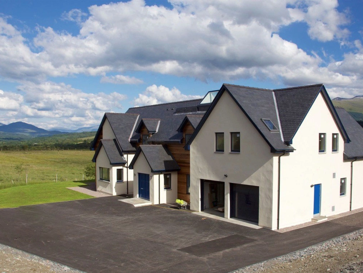 Modern two-story house with a mix of white and wooden exterior, situated in a scenic landscape. Lush green fields and mountains are visible in the background under a blue sky filled with fluffy clouds. A spacious driveway is in front of the house.