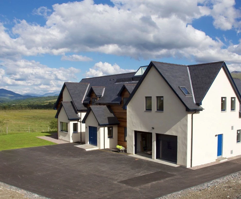 A modern two-story house with a sleek design, featuring a mix of white walls and wooden accents. Large windows and blue doors complement the exterior. The house is surrounded by a green lawn and open fields, under a bright blue sky with scattered clouds and distant mountains.