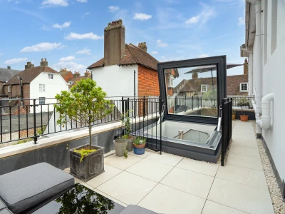 Modernes Balkon mit eleganten Kissenmöbeln und einem großflächigen Fenster, das auf die umliegenden Dächer einer Stadt blickt. Töpfe mit Pflanzen stehen auf dem Boden. Helle, freundliche Wolken am Himmel.