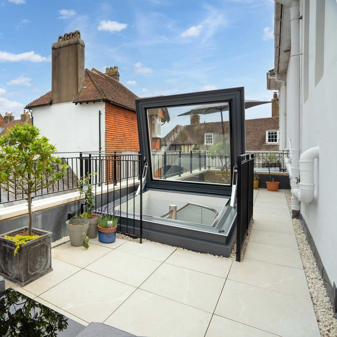 Modernes Balkon mit eleganten Kissenmöbeln und einem großflächigen Fenster, das auf die umliegenden Dächer einer Stadt blickt. Töpfe mit Pflanzen stehen auf dem Boden. Helle, freundliche Wolken am Himmel.