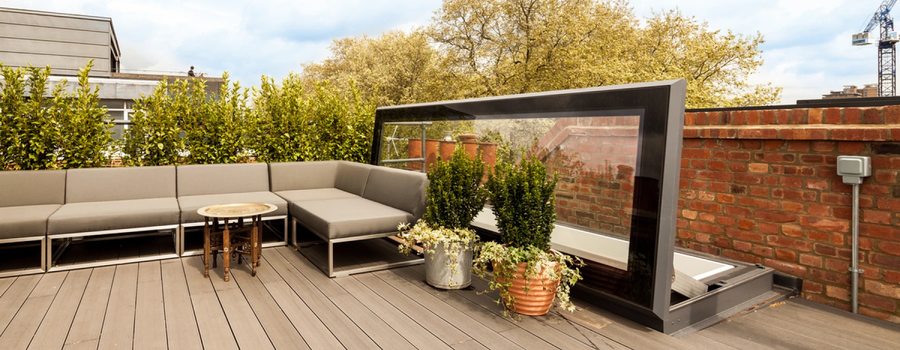 Terraza con un sofá gris en forma de L, una mesa circular y plantas en macetas. Al fondo, un panel de vidrio transparente y un cielo nublado.