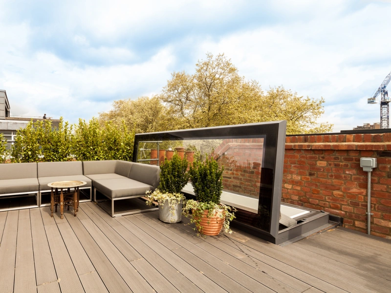 A stylish rooftop terrace featuring a modern gray sectional sofa, a small round table, and potted plants. The area is surrounded by greenery and includes a glass windbreak. Various buildings and a crane are visible in the background under a blue sky.