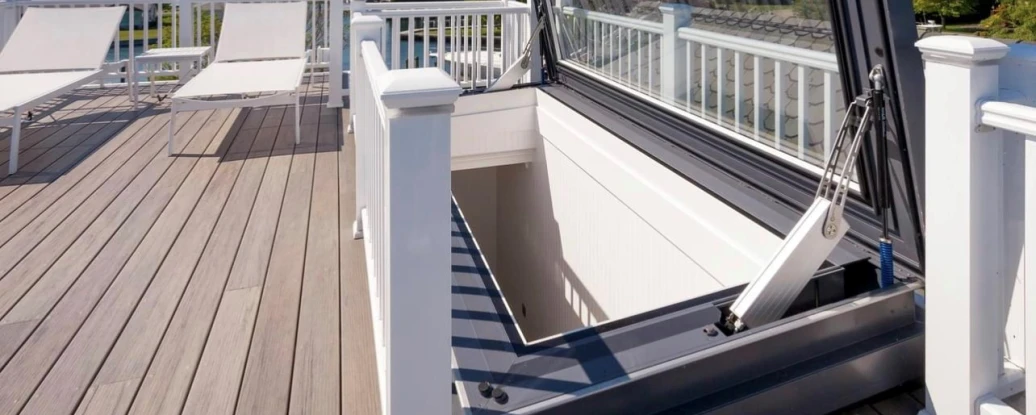 A rooftop deck featuring a large, open hatch that leads below. The surrounding area is adorned with white railings and two lounge chairs, with a scenic view of the sea and blue sky in the background.