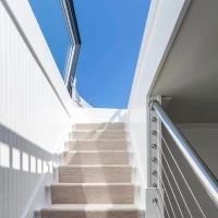 Escalera blanca iluminada por la luz del sol, con un pasamanos de metal y una alfombra beige que cubre los escalones. Al fondo, un cielo azul claro.