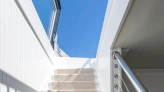 Escalera blanca iluminada por la luz del sol, con un pasamanos de metal y una alfombra beige que cubre los escalones. Al fondo, un cielo azul claro.