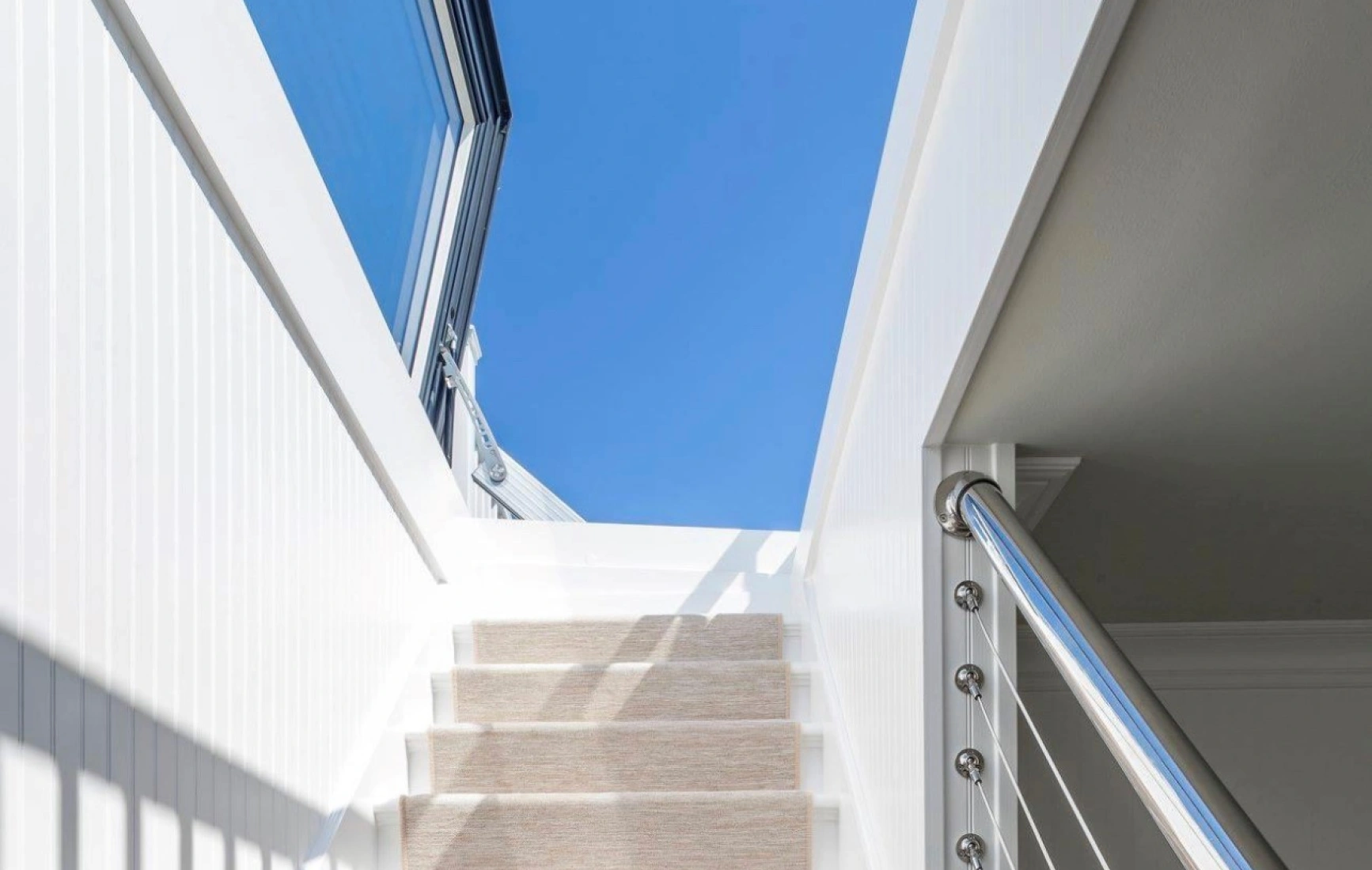 Un escalier blanc avec un tapis beige mène à un espace lumineux sous un ciel bleu. La main courante en métal ajoute une touche moderne.