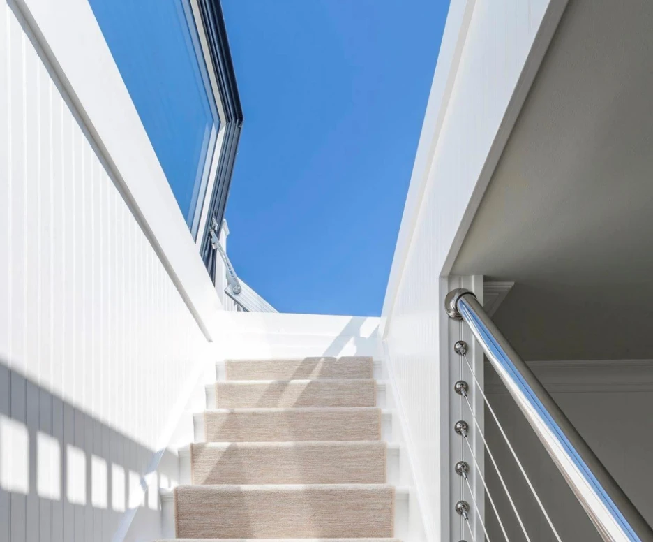 A well-lit staircase with soft beige carpet leading upward, flanked by sleek white walls and a modern railing. The bright blue sky is visible through large windows at the top.