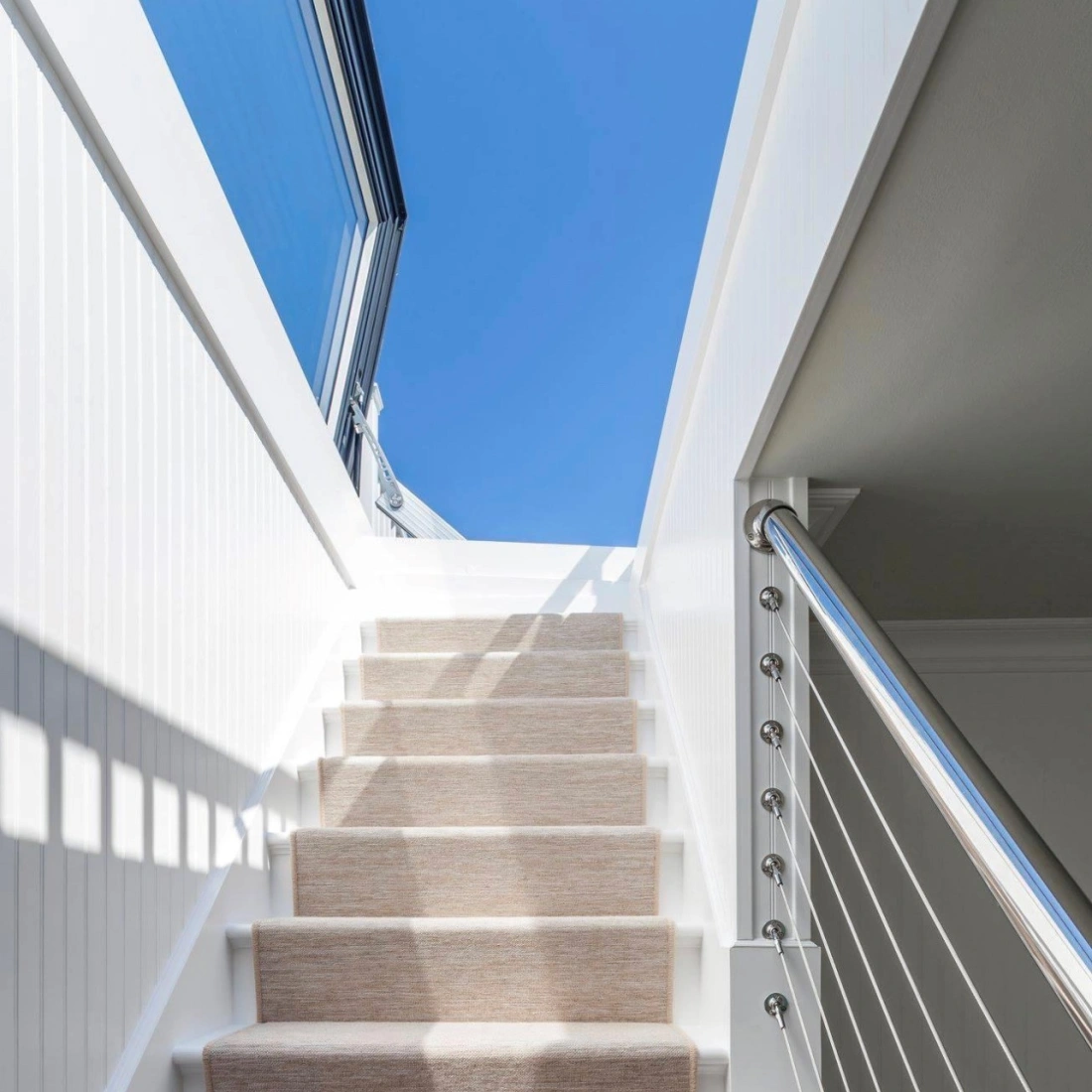 Staircase with white walls and a light-colored carpet runner leading up to a bright blue sky visible through a large window. Modern railing with stainless steel accents.