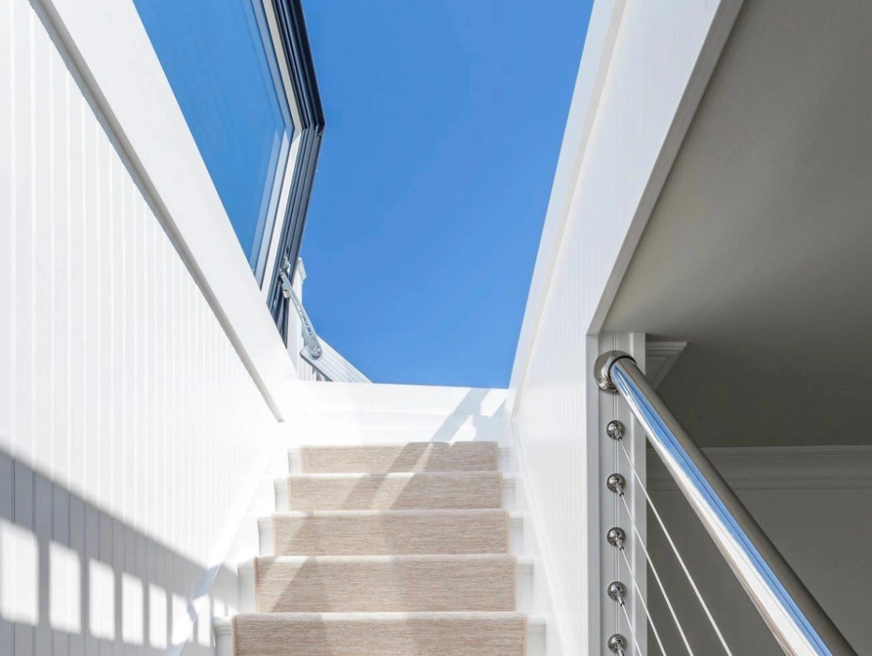 Staircase with white walls and a light-colored carpet runner leading up to a bright blue sky visible through a large window. Modern railing with stainless steel accents.