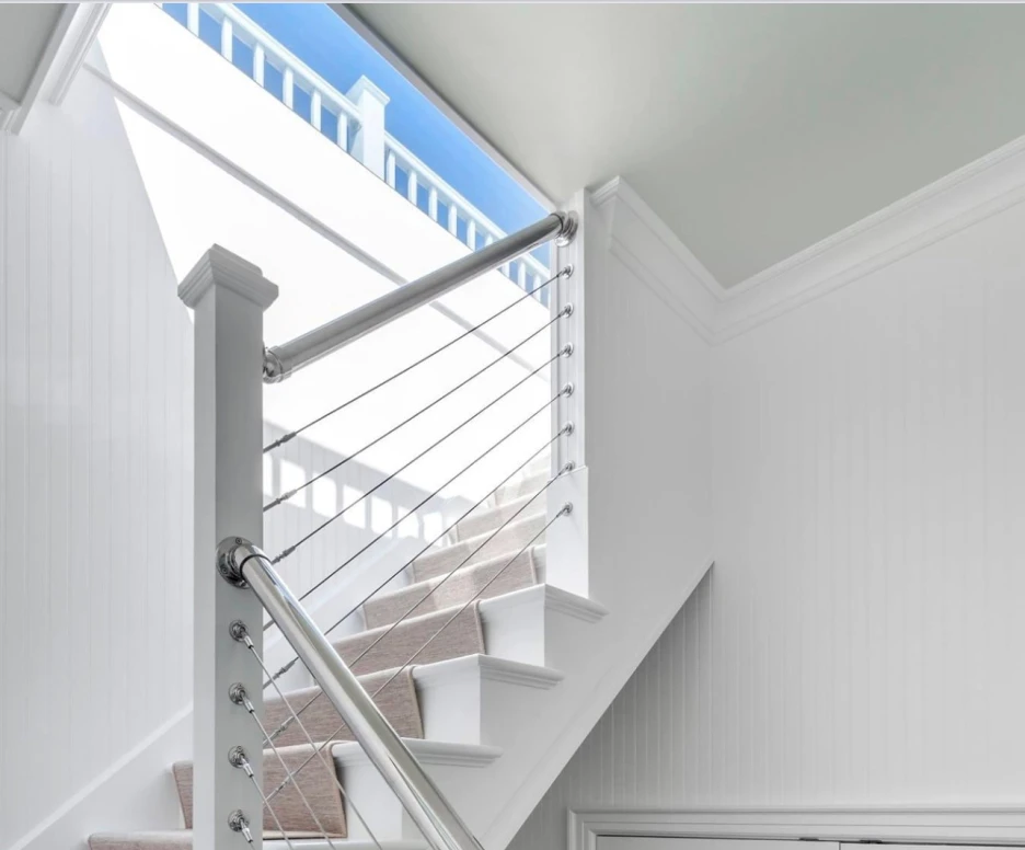 A modern staircase with a sleek metal railing and wooden steps. Natural light streams in from a large window at the top, illuminating the white-paneled walls. A framed picture hangs on the wall near the bottom of the stairs.