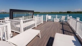 Terraza con suelos de madera, tumbonas blancas y barandillas blancas, frente a un lago. Se observa un cielo despejado y un paisaje verde en el horizonte.