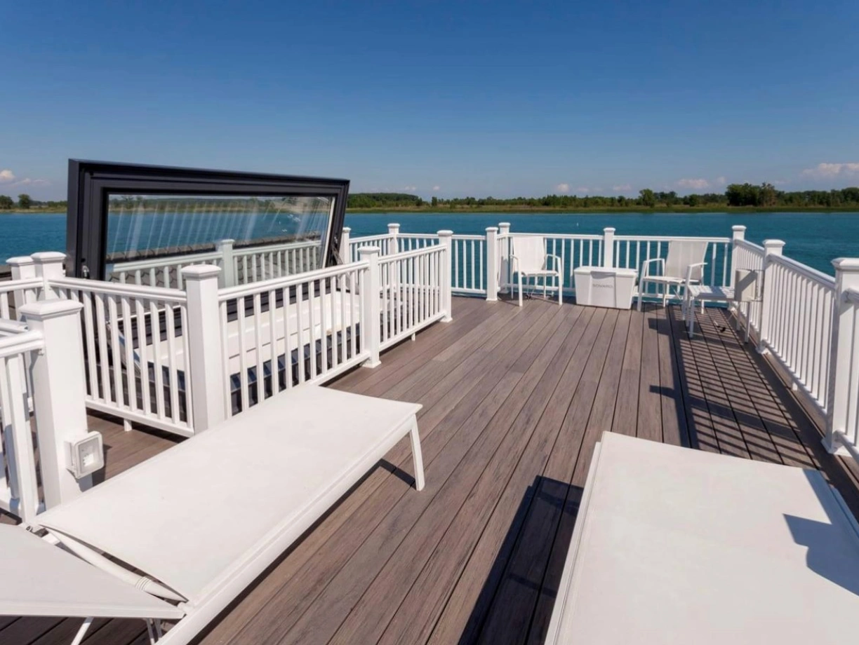A spacious wooden deck with white lounge chairs and railings overlooks a calm blue body of water, under a clear blue sky.