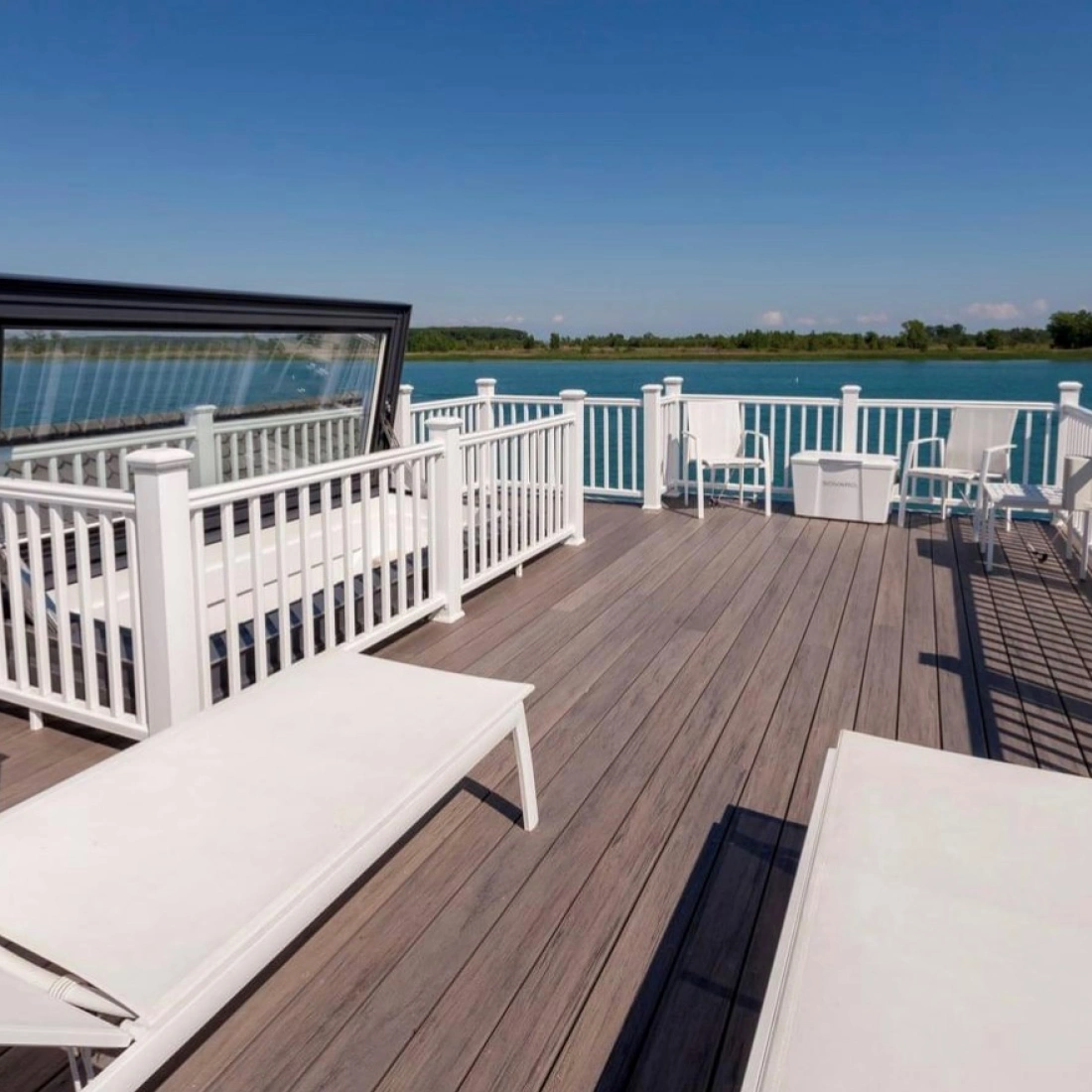 A spacious waterfront deck features white lounge chairs and railings, overlooking a calm blue lake under a clear blue sky.