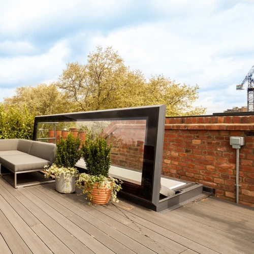 A stylish rooftop patio with gray seating, a round table, and potted plants. The space is surrounded by greenery and features a modern glass partition. Brick walls and a blue sky create a serene atmosphere.