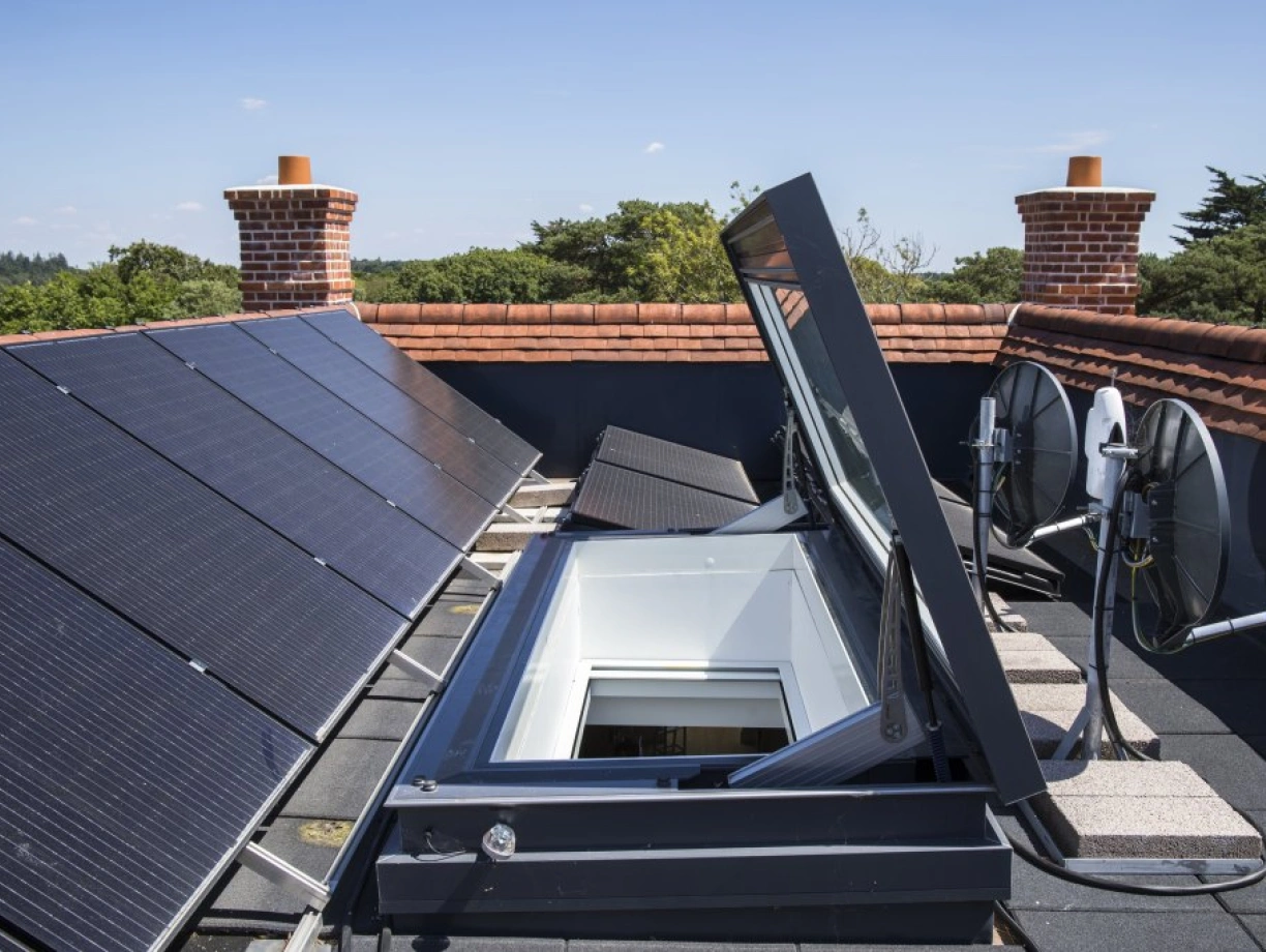 A rooftop scene featuring solar panels, a skylight opening, and two satellite dishes. The background shows trees and a clear blue sky.