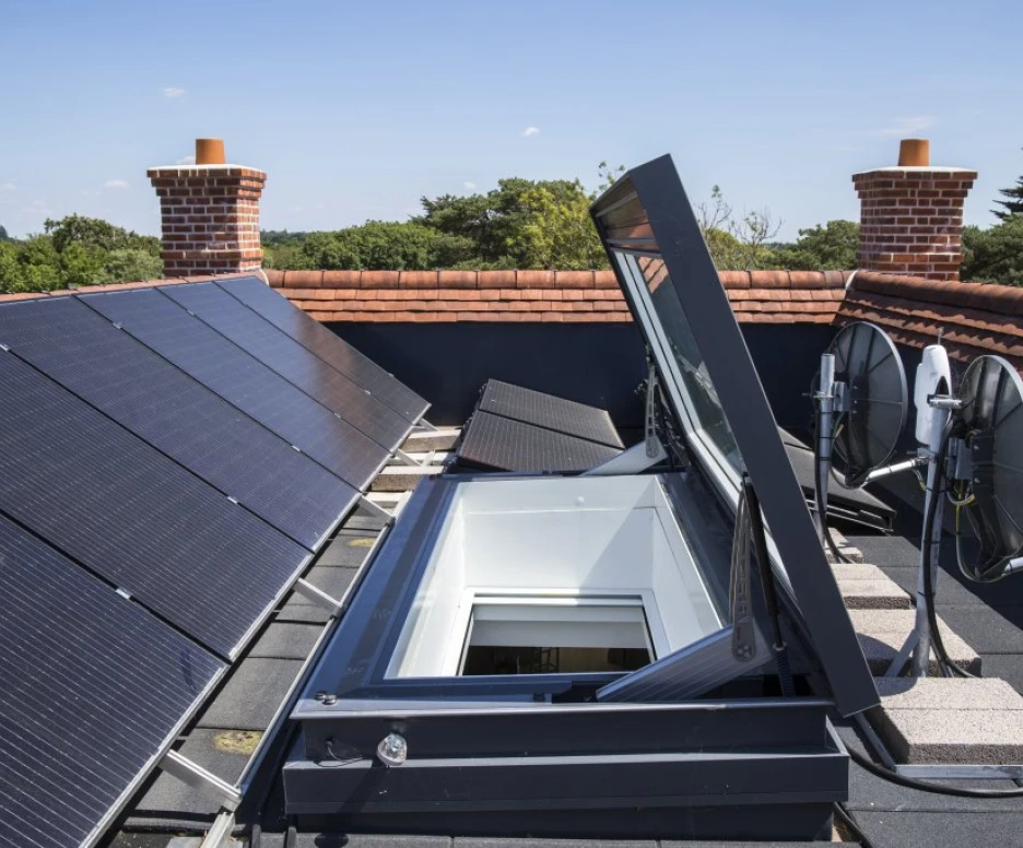 Panneaux solaires installés sur un toit, avec un puits de lumière ouvert et des antennes paraboliques à côté. Le ciel est clair et des arbres verdoyants se dressent à l'arrière-plan.