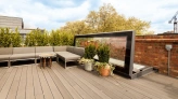 A stylish rooftop patio with gray seating, a round table, and potted plants. The space is surrounded by greenery and features a modern glass partition. Brick walls and a blue sky create a serene atmosphere.