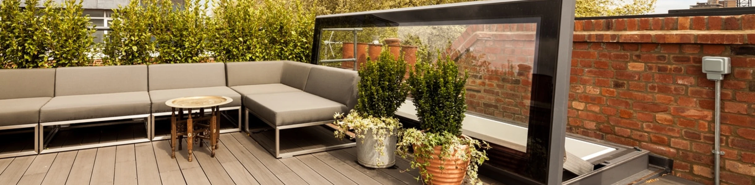 Une terrasse moderne avec un canapé gris, une table ronde, et des pots de plantes. En arrière-plan, des arbres feuillus et un mur en briques. Le ciel est partiellement nuageux.